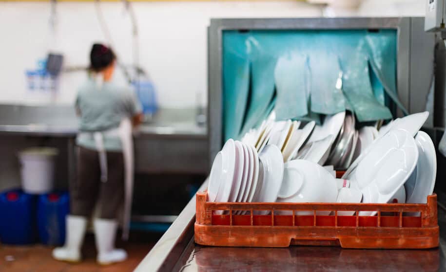 The right-side features clean dishes emerging from a restaurant's industrial kitchen dishwasher. At the same time, a blurry employee in the back washes dishes in a sink using a commercial boiler heating system to meet sanitation requirements.