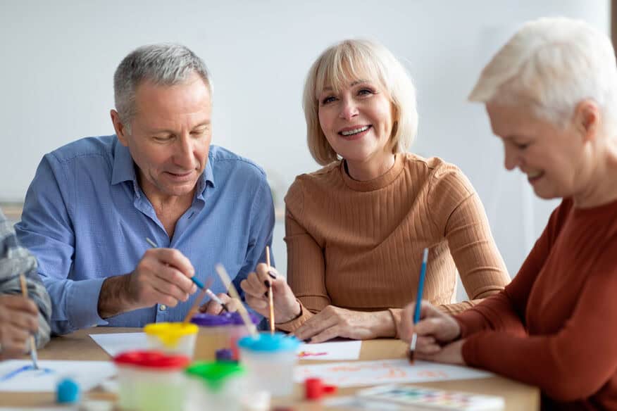 Three older adults sit around a table full of art supplies. They smile as they paint during art therapy for seniors.