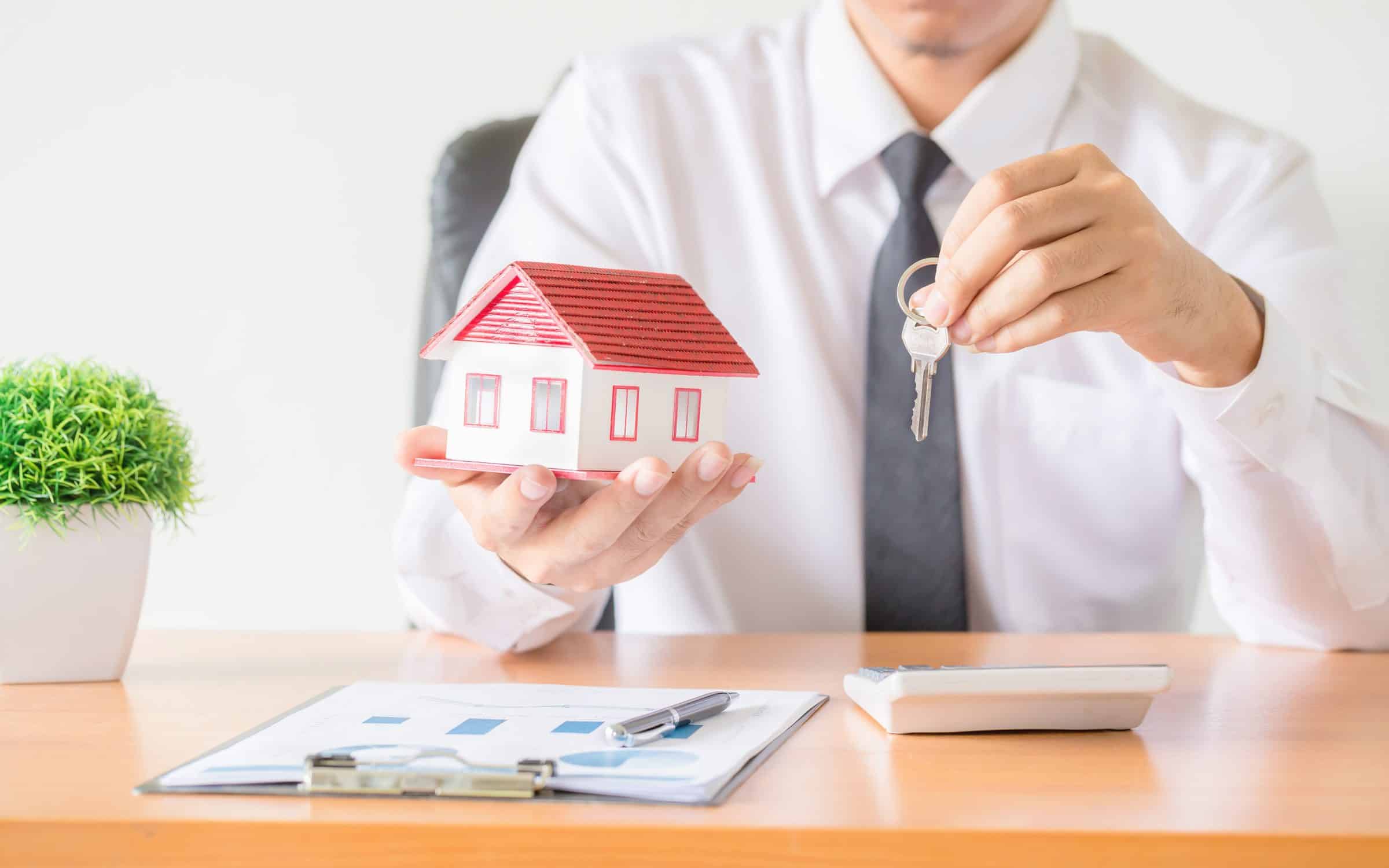 A businessman in a white shirt and tie sits behind a desk. He is holding a small model house and keys.