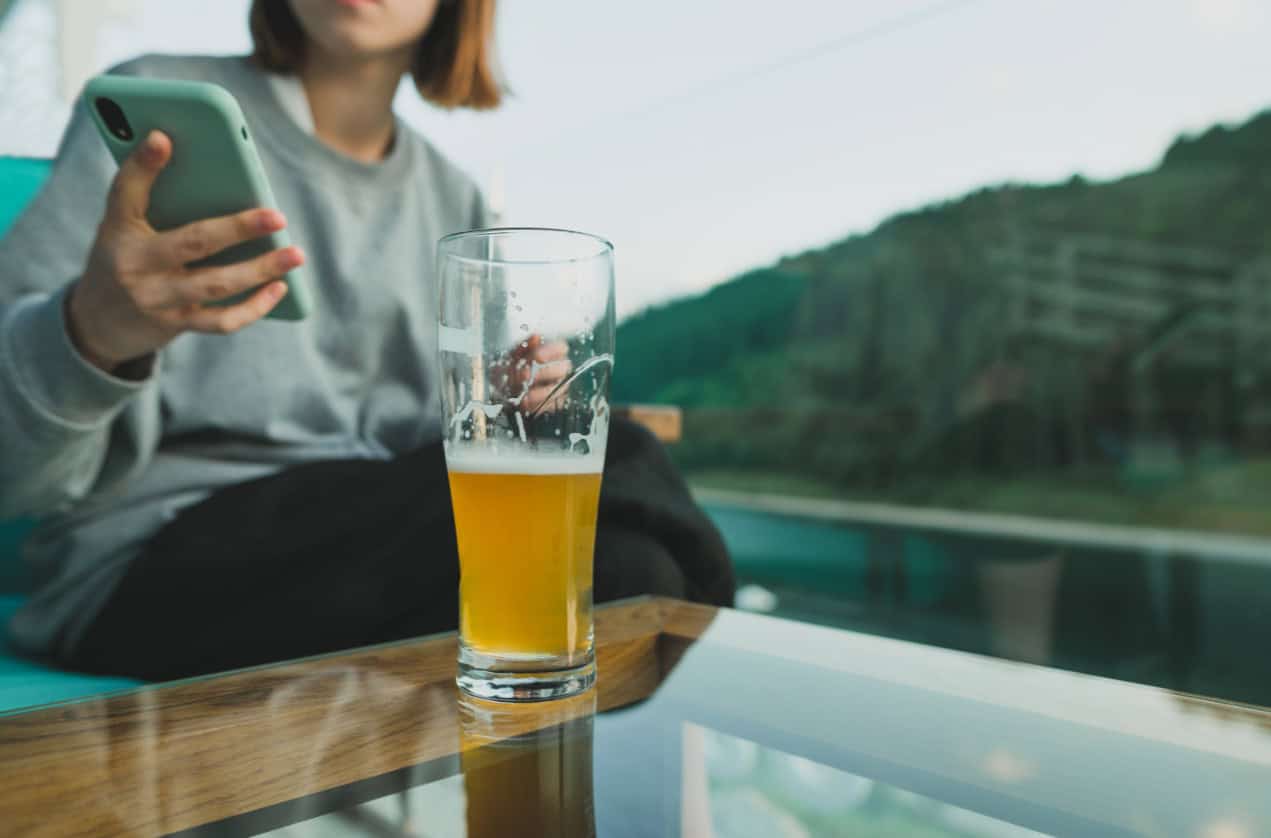 A pint glass, half full of beer, sits on a table outside. A woman sits behind the glass, holding her phone.