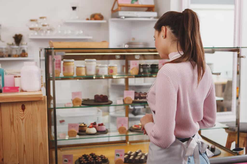 A baker wearing an apron examines the cupcakes and baked goods in a bakery case.