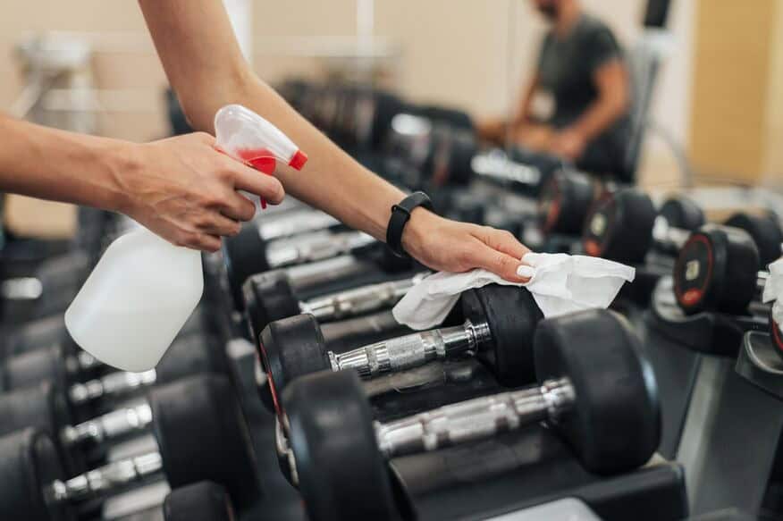 A female gym employee uses a spray bottle and a cloth to carry out gym cleaning best practices.