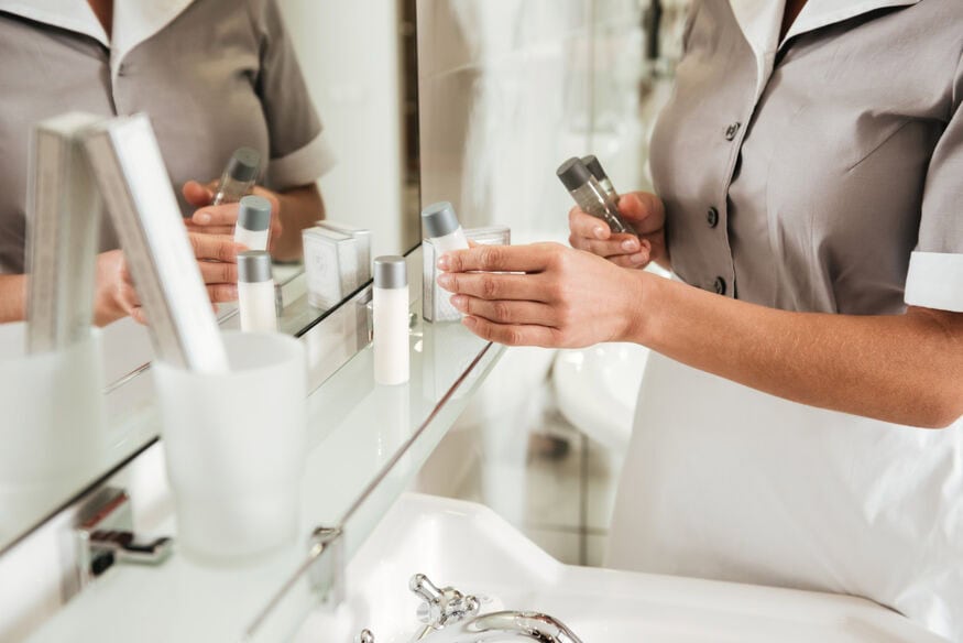 A female hotel housekeeper in a gray uniform restocks single-use shampoo and conditioner on the shelf in front of the bathroom mirror.