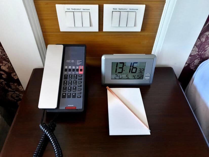 A hotel phone, a large digital alarm, and a pencil and pad of paper are on a desk. On the wall behind them are electrical switches.
