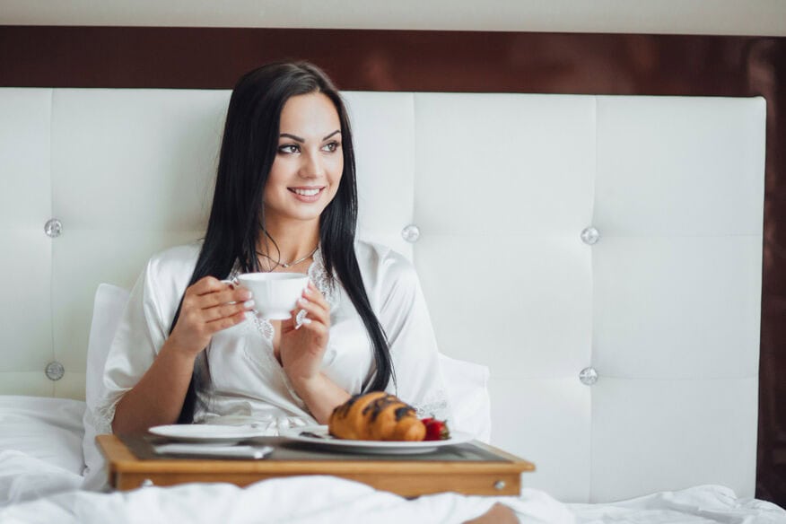 A woman sits on a hotel bed enjoying coffee and room service, one of many amenities hotels offer. 