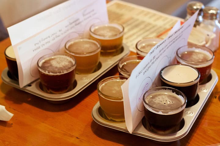 Two beer flights, each with 6 glasses of beer separated in muffin tins, sit on a table with sheets naming all the varieties of beer.