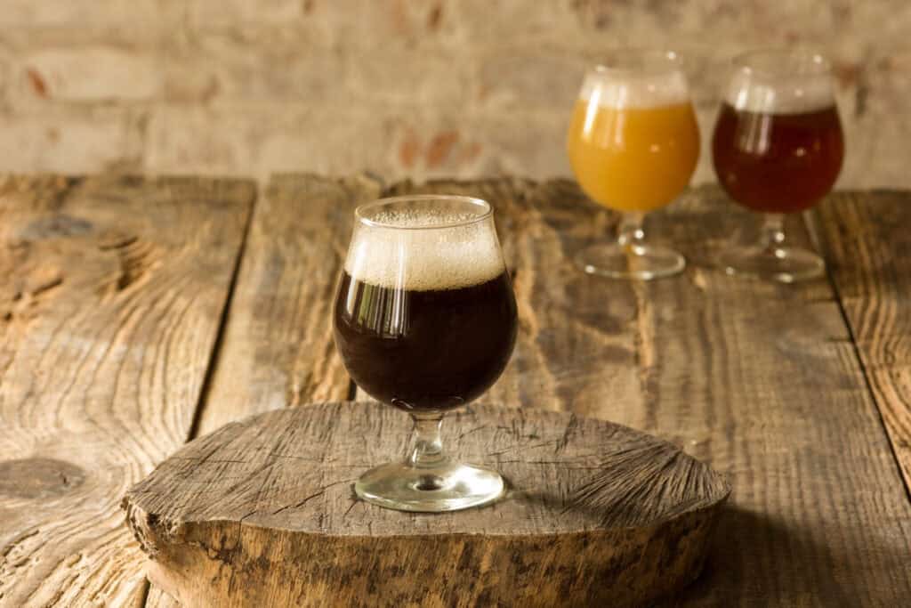 Three glasses of beer, each of a different color, sit on a wood table. Two glasses sit in the background and one glass sits on a wood slice front and center.