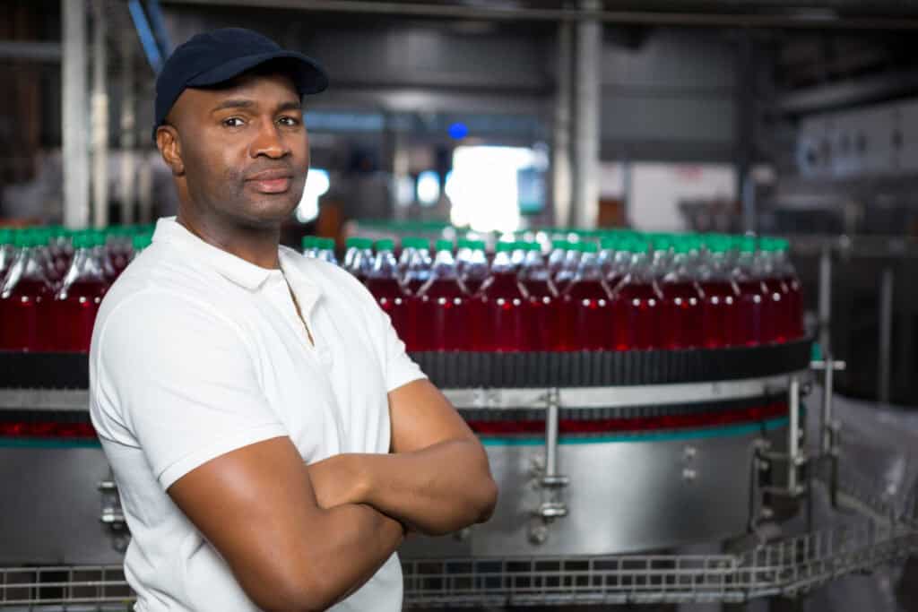 An African American man wearing a white shirt and black hat stands confidently with his arms crossed in front of a manufacturing conveyor belt packed with bottles of liquids.