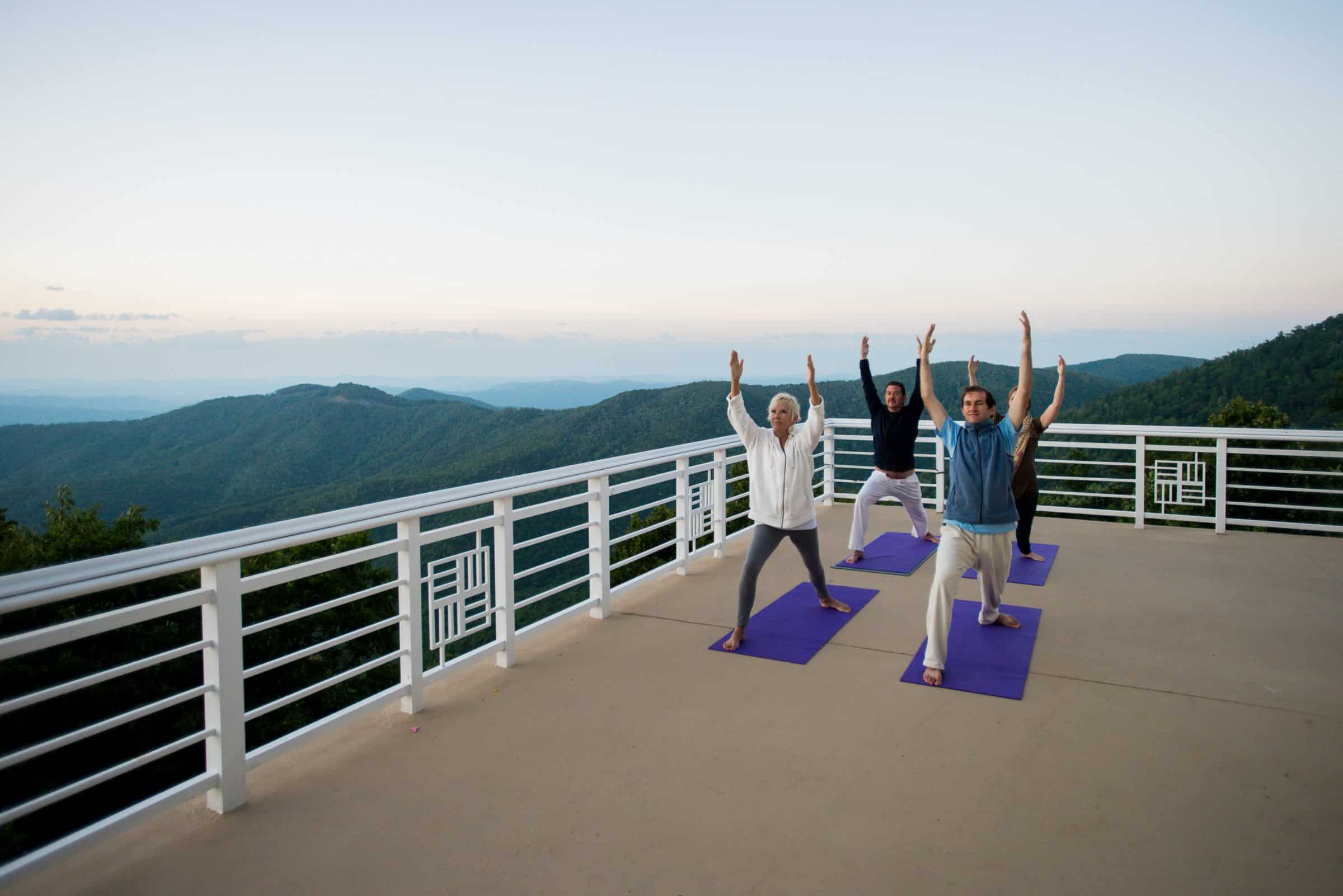 A group of men and women do yoga on a patio overlooking a vast landscape of mountains and blue sky.