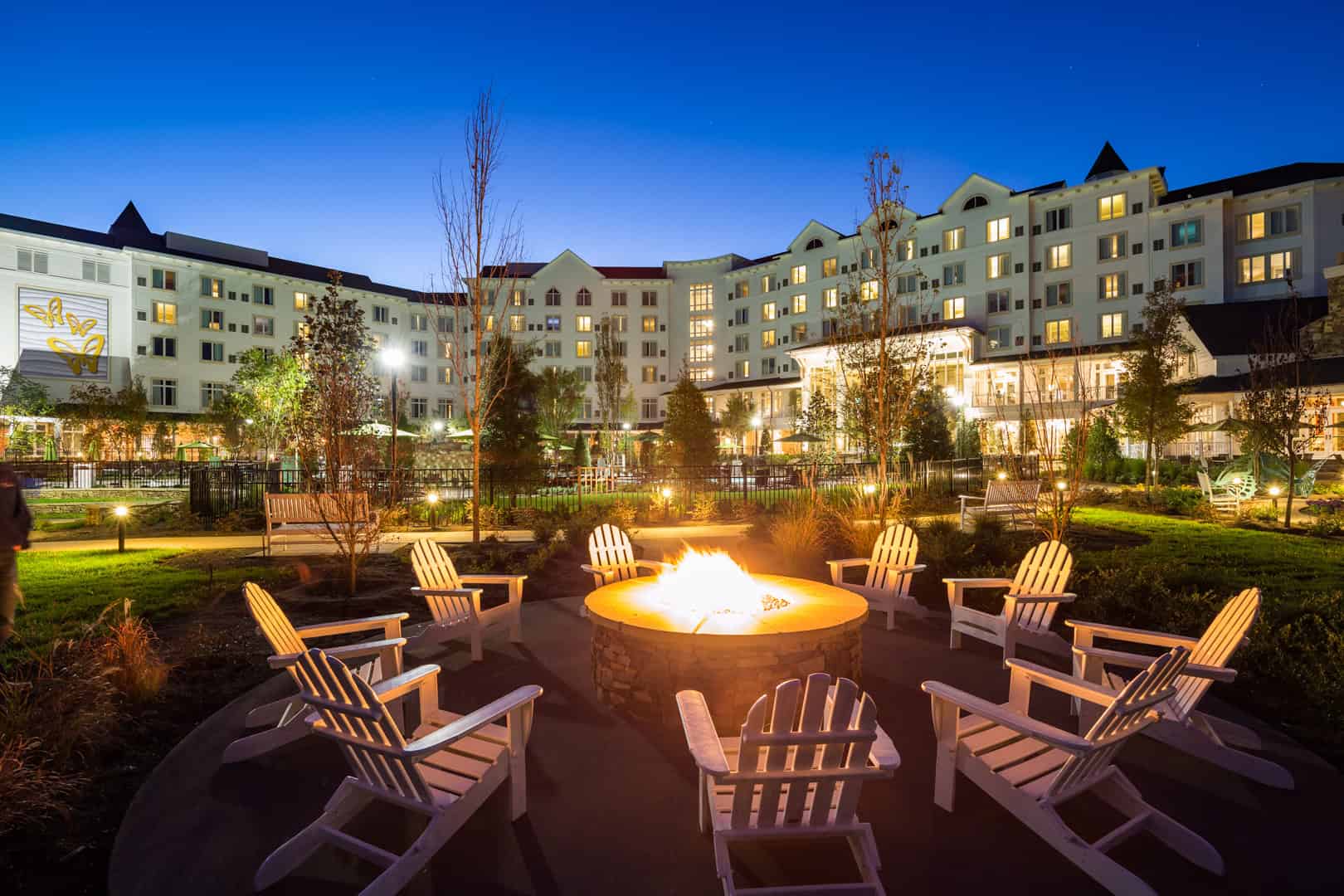 A broad shot of Dollywood Resort's courtyard at dusk, with a view of the whole resort, landscaping, and Adirondack chairs around a lit bonfire pit.
