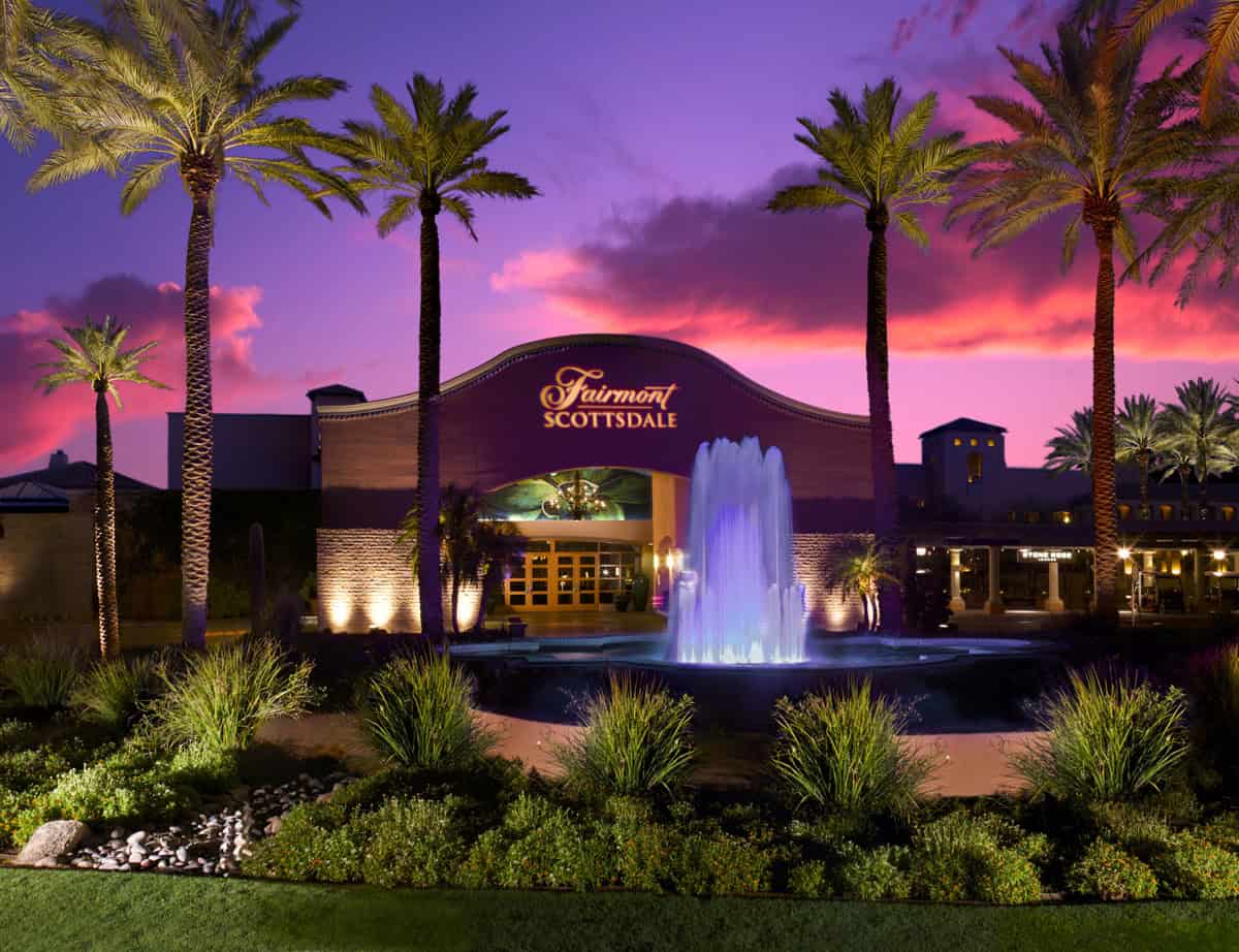 The front facade of the Fairmount Scottsdale resort and water water fountain, surrounded by lush landscaping, palm trees, and a pink and indigo sky at sunset.