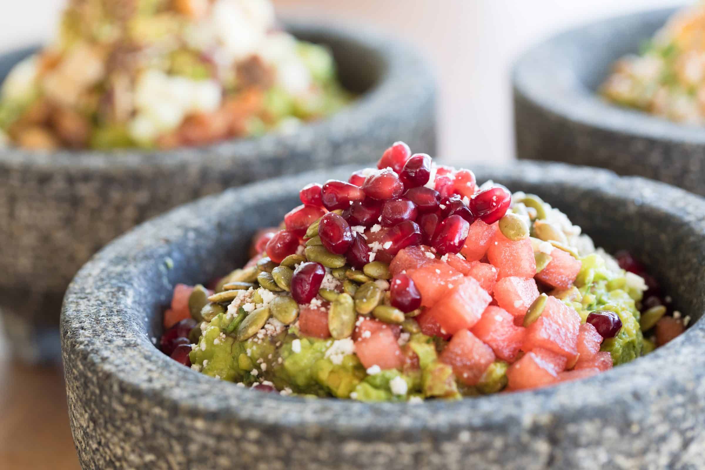A stone bowl full of house-made guacamole topped with tomatoes, pine nuts, and pomegranate seeds.