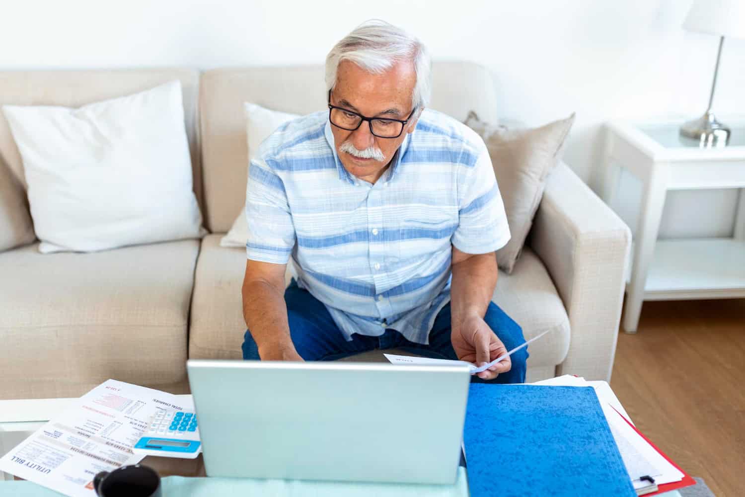 An older adult man sits on a couch, working at a laptop, with paperwork and folders spread out on the table in front of him.