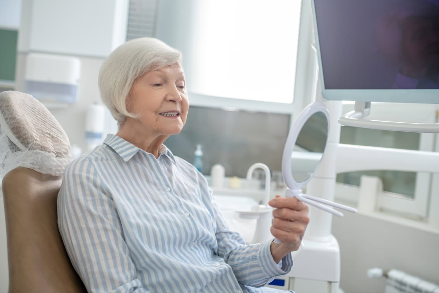 An older woman with white hair, wearing a blue and white striped shirt, sits in a dentist chair and holds a mirror up to her face, smiling and admiring her teeth.