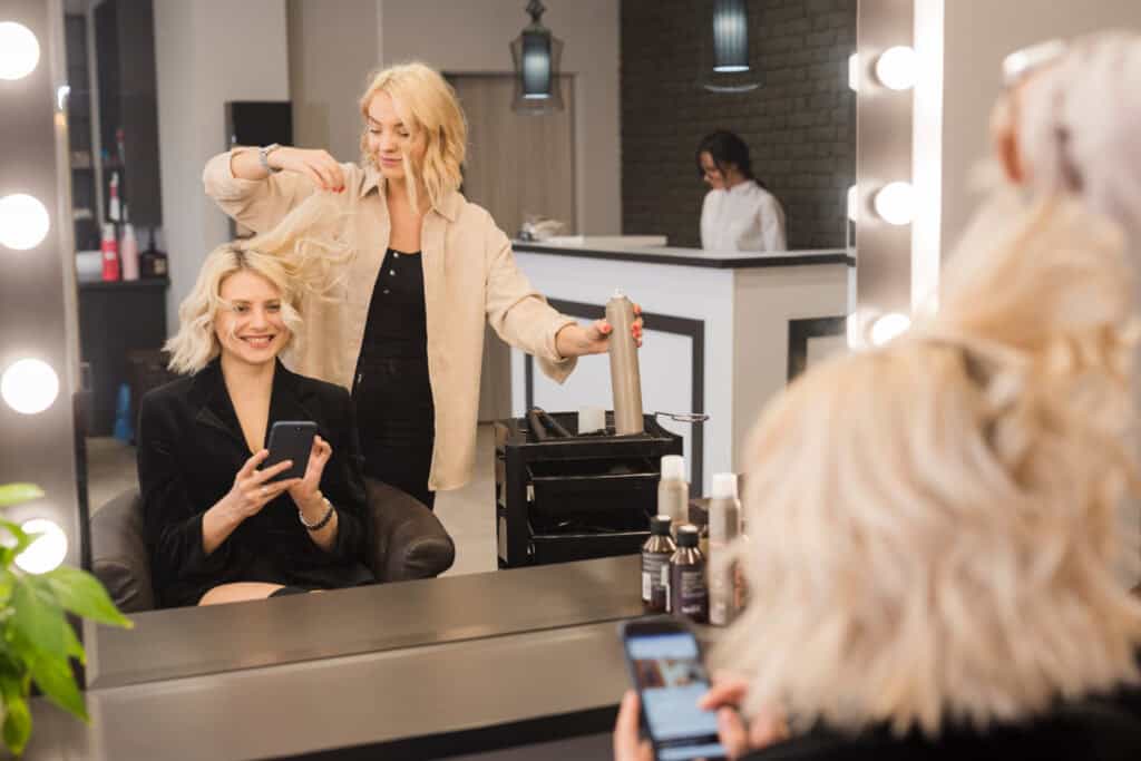 A woman sits in a salon chair, smiling into the mirror as a hair stylist prepares to spray and style her hair.