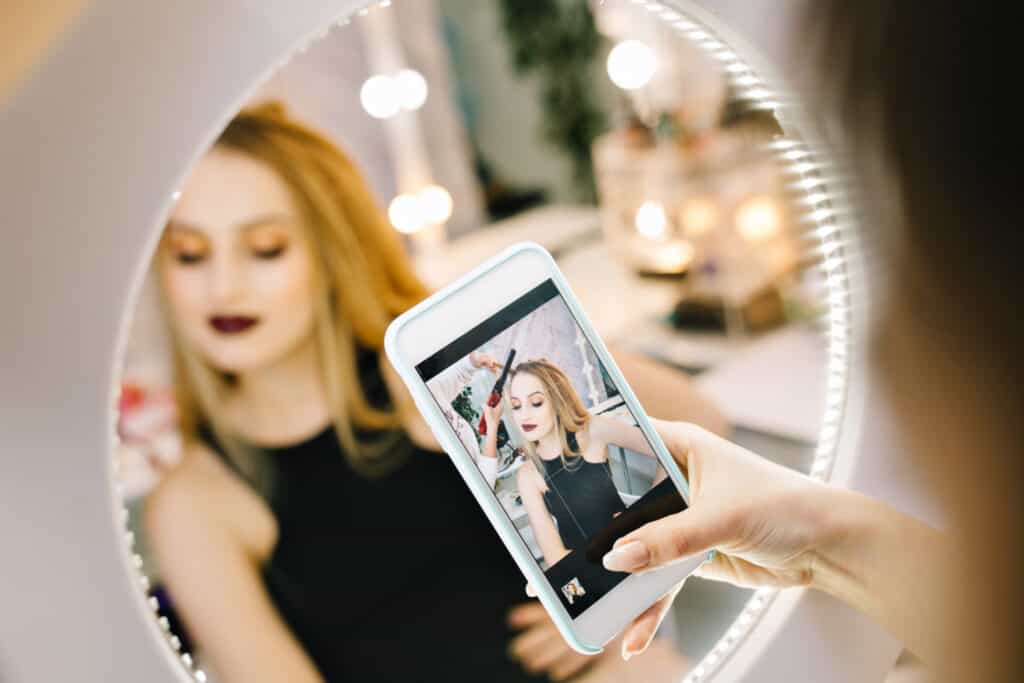 A woman’s hand holds a phone camera up to a mirror. The woman’s reflection, which shows her getting her hair styled, is seen on the phone’s screen.