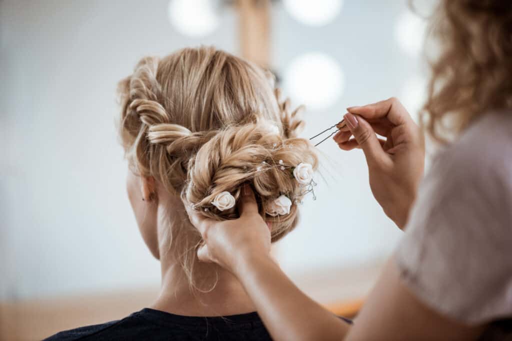 A hair stylist adds a pin to an elaborately braided updo, with small flowers arranged in her client's hairstyle.