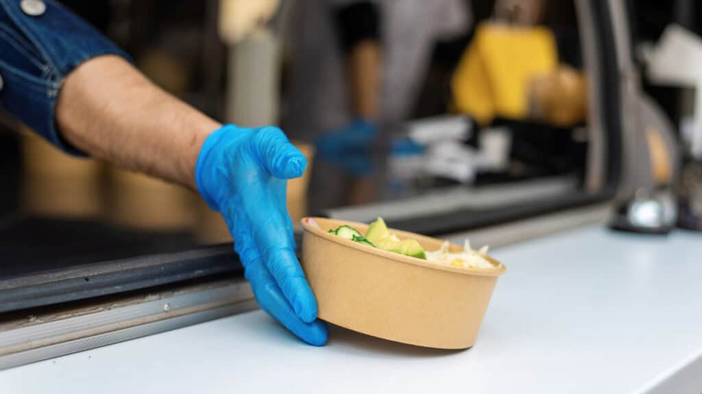A man wearing a blue latex glove places a brown paper bowl filled with salad on a white countertop.