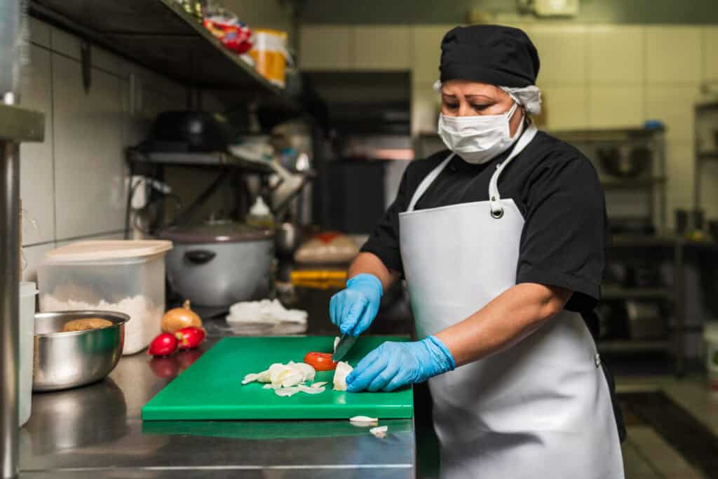 A cook in a commercial kitchen wears blue laytex gloves as she slices vegetables on cutting board. She's wearing a black chef's outfit, white apron, and a white face mask.