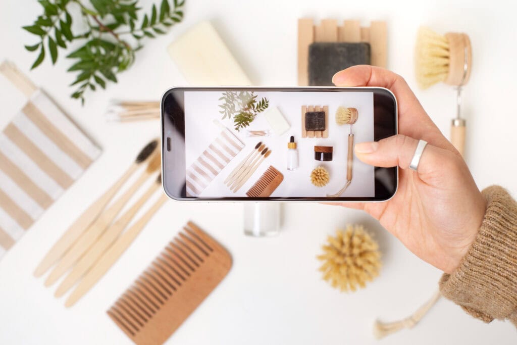 A selection of beauty opbjects - makeup, brushes, makeup bags, and more, lay flat on a white backdrop. A woman holds her phone over the setup, taking an aesthetically pleasing photo of the beauty spread for social media.