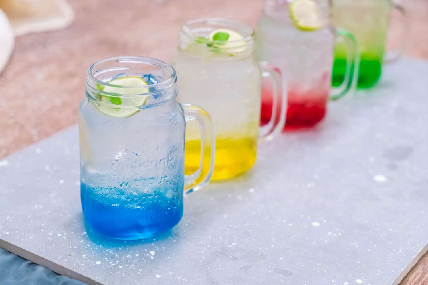 Four mason jars filled with colorful blue, yellow, blue, and green infused waters sit in a row on a stone slab. Each mug has a garnish of lime and mint.