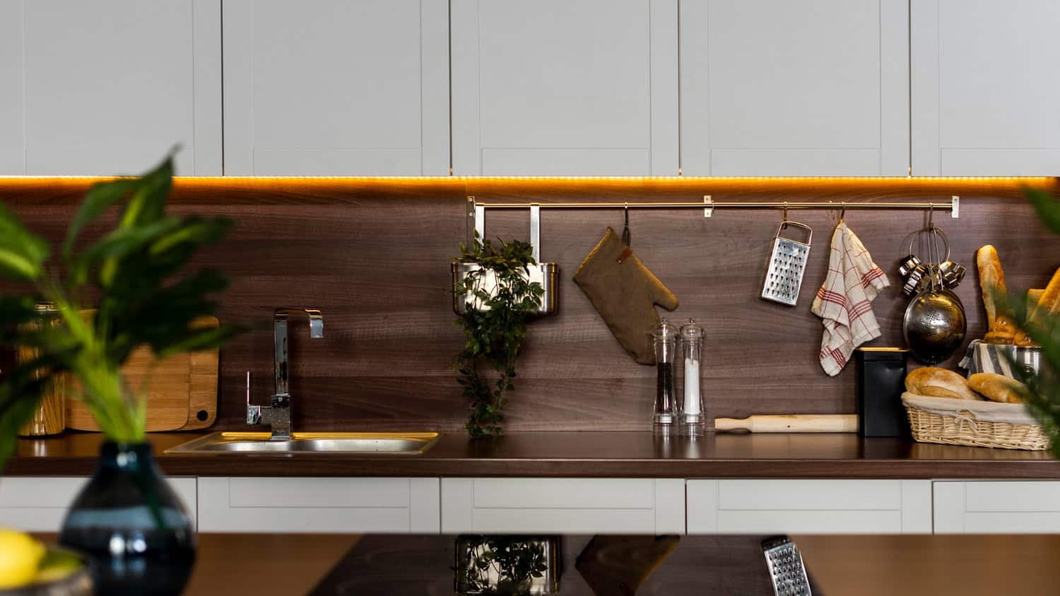 A modern kitchen has white wood cabinets and dark wood countertops and backsplash. The cabinets have soft under cabinet lighting giving off a warm amber glow and illuminating various kitchen accessories.