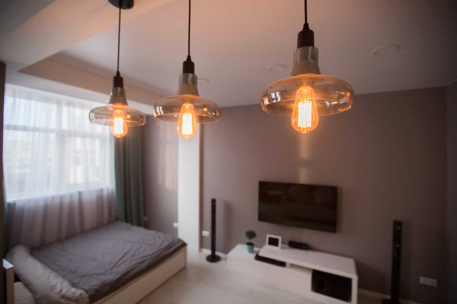 Three small pendant lights with soft amber-glowing Edison light bulbs hang from a ceiling in a dark bedroom.