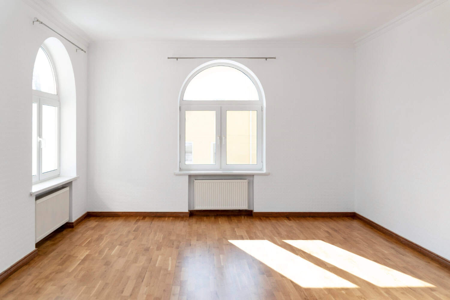 A small apartment with natural hardwood floors and white walls has two arched windowsills with double windows on opposing walls. Natural light floods in from one of the windows, casting two bright strips of light onto the floor.