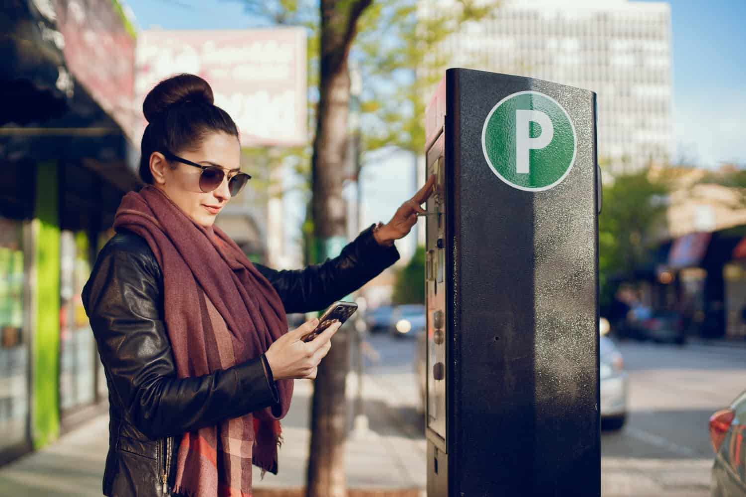 A young woman stands at a parking payment box, marked with a white letter P in a green circle. She's holding a smart phone and inserting her card into the payment box.