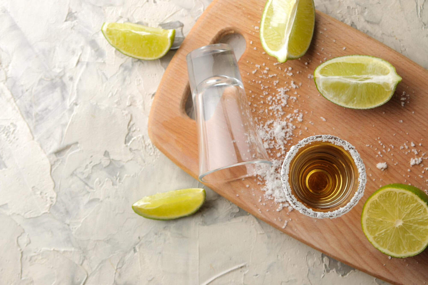 Shot glasses sit on a small wooden cutting board surrounded by lime wedges and loose salt.