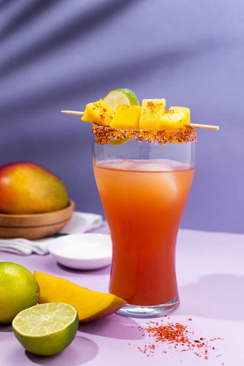 A Michelada cocktail served in a pint class, garnished with a spicy rim and mango and lime slices.