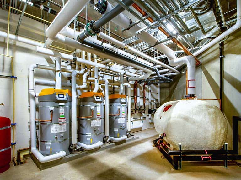 Several large commercial water heaters are installed against a wall, with pipes leading up to the ceiling.