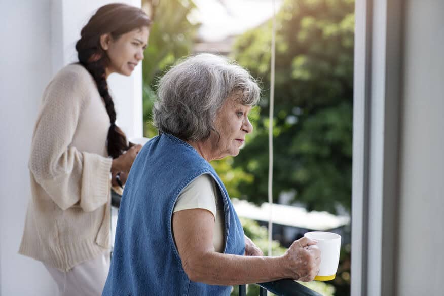 An elderly woman and a younger woman drink coffee together while looking out a large senior living facility window. 