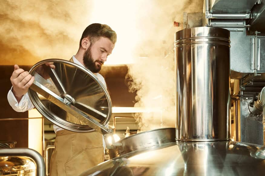 A bearded man opens the lid on a stainless brewing vat as steam rises behind him. Brewery boilers are essential to the production of craft brews.