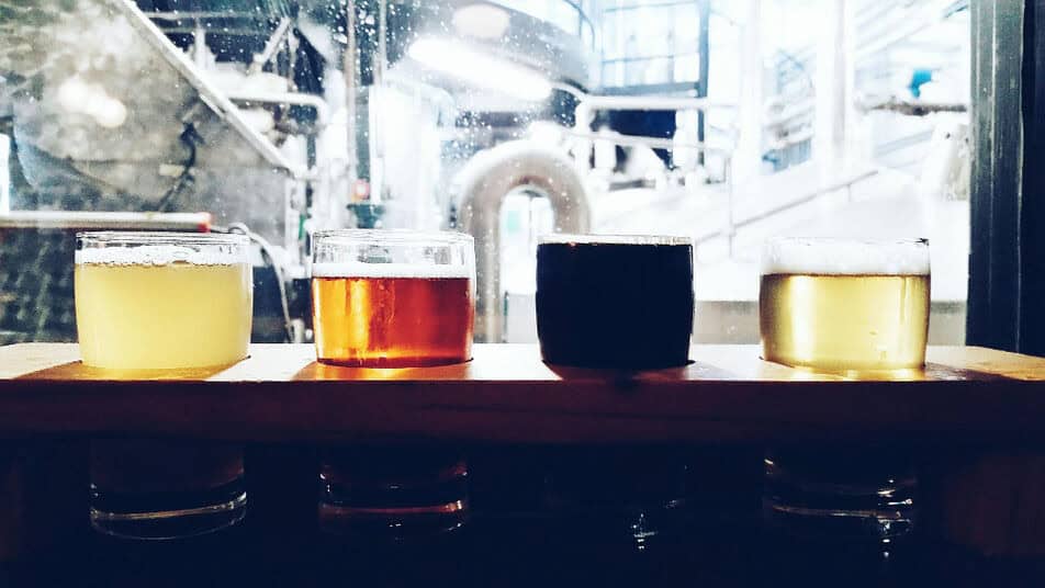 Four different types of craft brews in glasses are ready for tasting. Industrial brewing equipment, like a brewery boiler, are in the background.