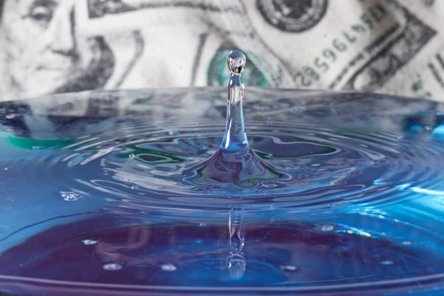 A close-up of a drop of water into a glass of water, with US dollars out of focus in the background.