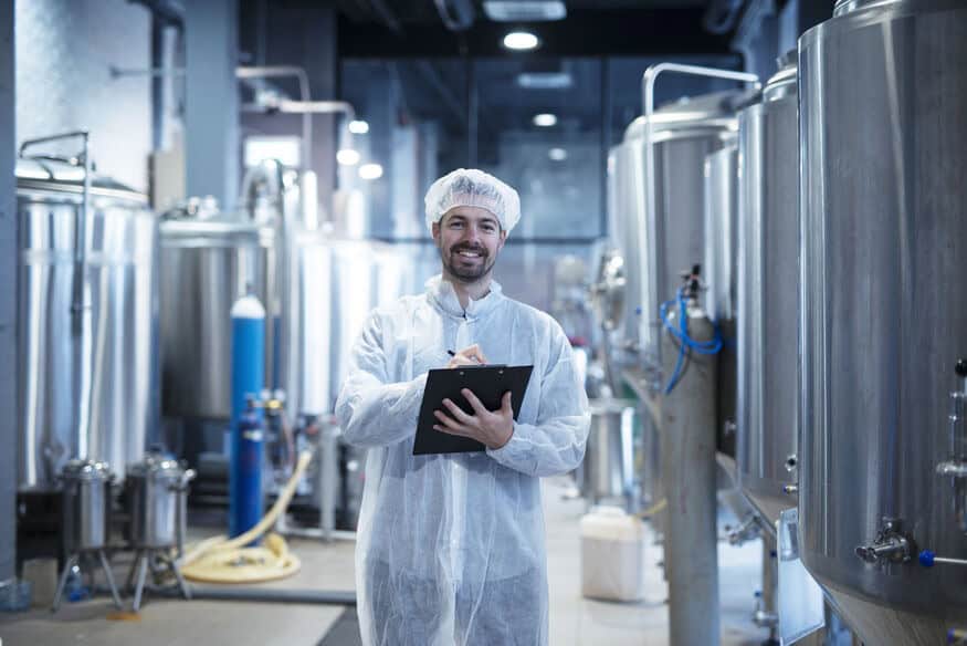 A smiling technician holds a clipboard while standing in the aisle lined by large stainless-steel vats used as an energy-efficient solution in food manufacturing.