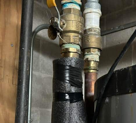 Insulation is taped to the hot water pipes in the corner of a cinder block room.