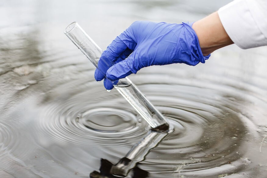 A blue-gloved hand holds a test tube of groundwater taken from the pool of water underneath. Hard water is one reason water softeners for breweries have become a necessity.
