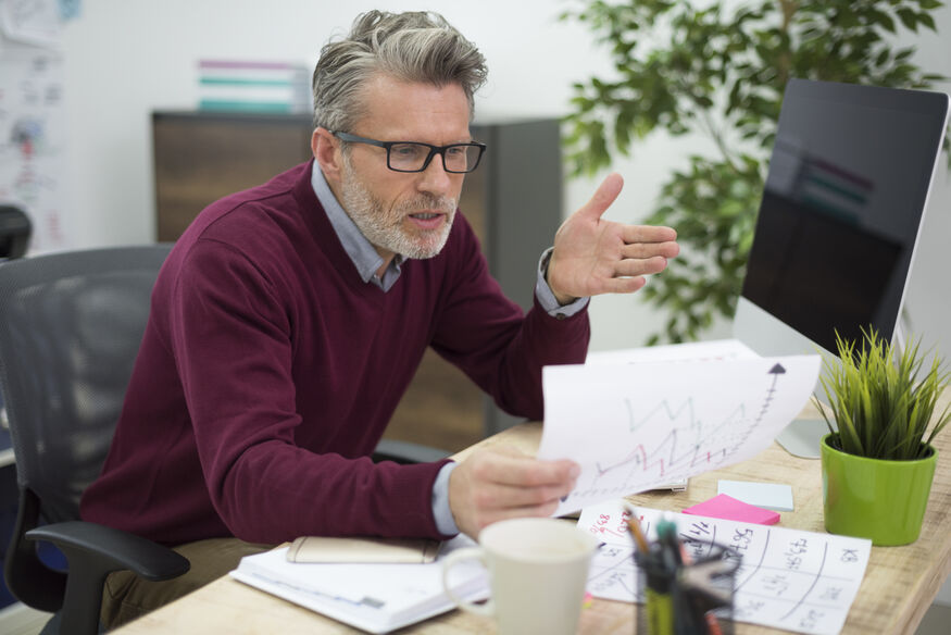 A man with glasses sits behind an office desk, looking confused and dumbfounded at the energy bill in his hands, which is higher than expected because of a low boiler efficiency rating.
