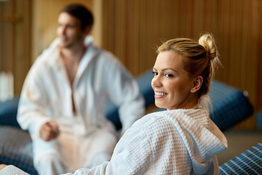 A woman in the foreground and a man in the background, both wearing white robes, wait for their spa treatments. In the debate between boilers vs water heaters, hotels consider the quantity of hot water required. 