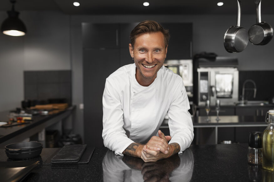 A smiling chef leans on a shiny stainless-steel counter, in front of an immaculate kitchen cleaned using hot water.