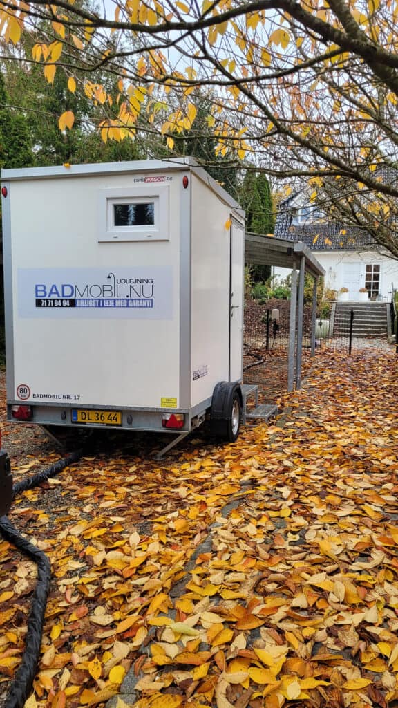 Kompakt toiletvogn placeret på efterårsblade ved hus.