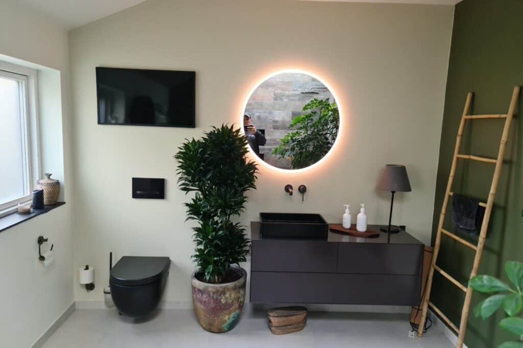 Lyst badeværelse med sort væghængt toilet, mørkt vaskemøbel, et belyst rundt spejl og en stor grøn stueplante.