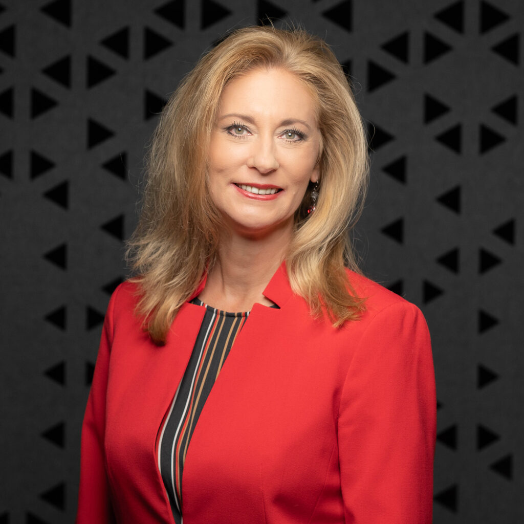 A professional portrait of business leadership consultant Diane Welhouse, Owner of Welhouse Consulting, wearing a professional red blazer in front of a black background.