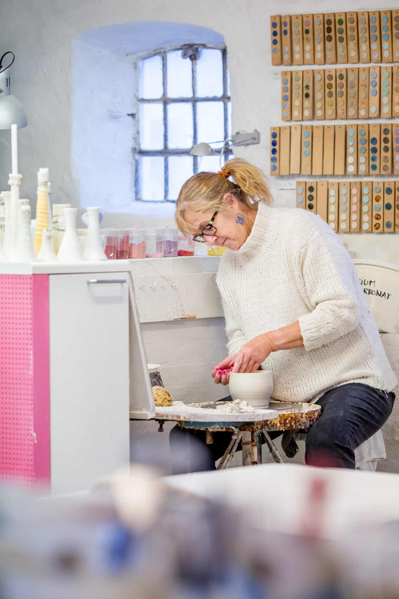 Kvinde arbejder med keramik i værkstedet, omgivet af værktøjer og keramik.