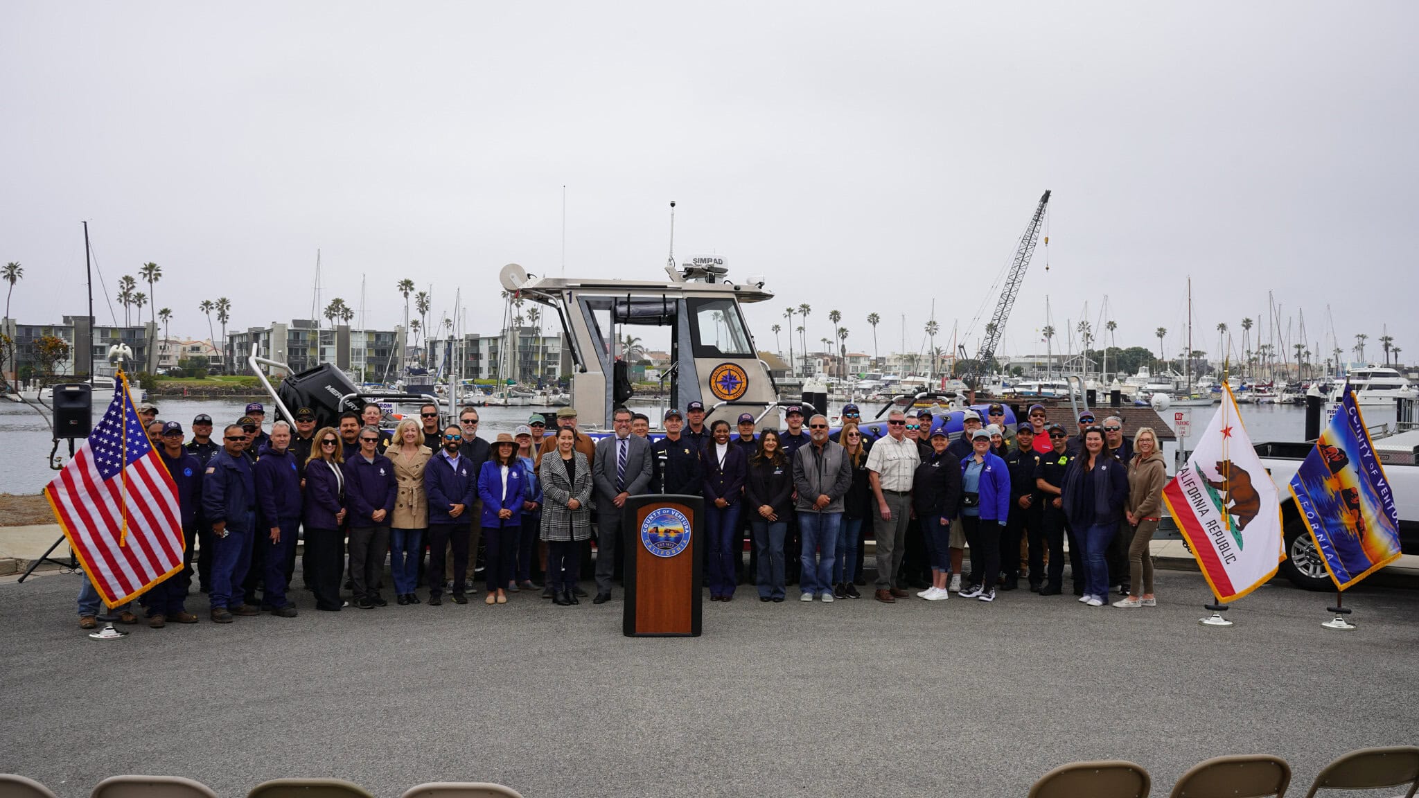 A large group of people stands behind a podium with official seals, flanked by U.S. and California flags, in front of a docked boat at a marina.
