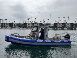 A blue and gray Harbor Patrol boat with a uniformed officer onboard moves through the water near a marina with palm trees and buildings in the background.