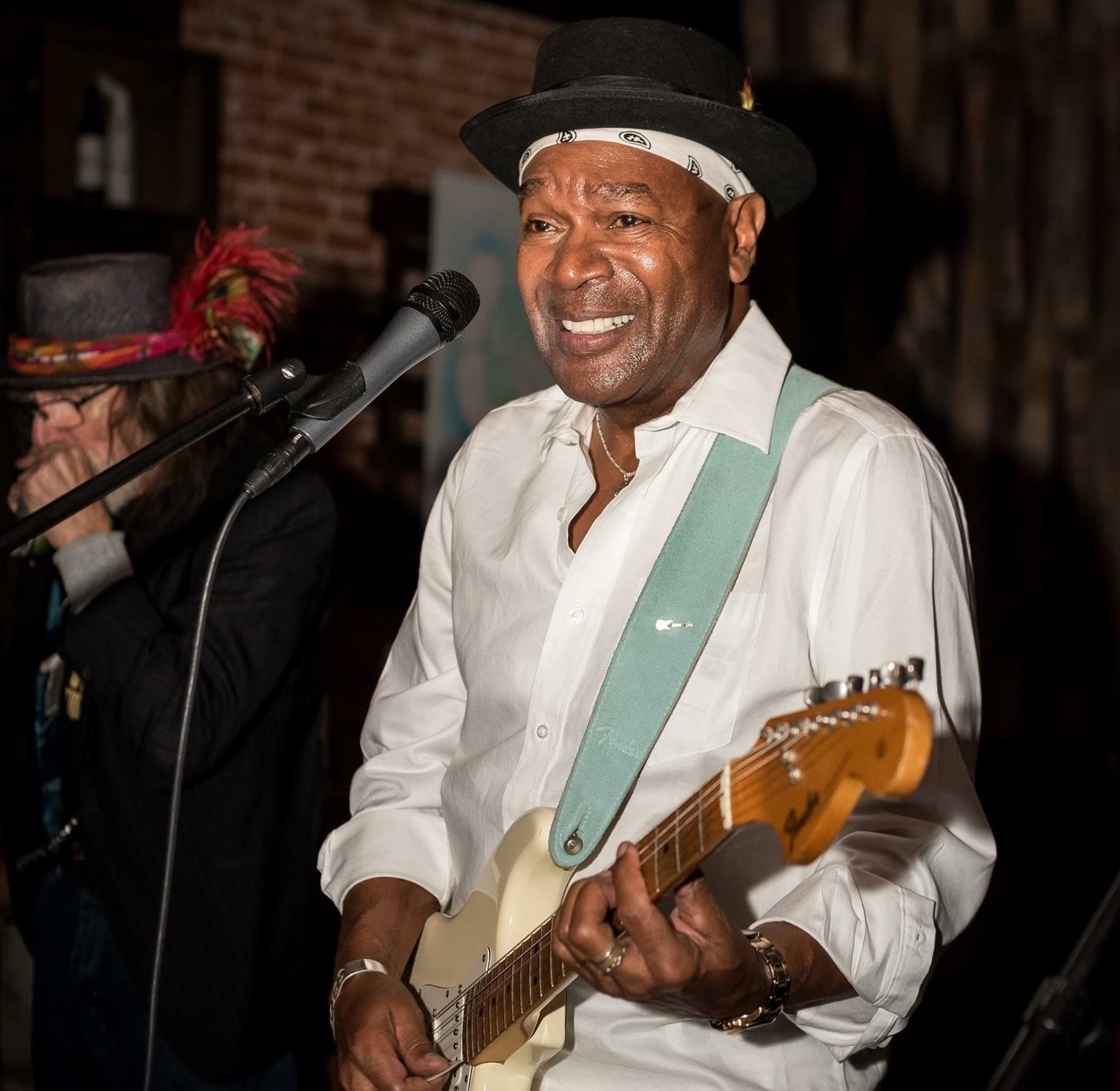 A man wearing a white shirt and black hat plays an electric guitar and sings into a microphone; another musician is in the background.