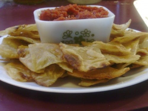 Image shows a white plate with homemade tortilla chips piled on it. At the center is a small white dish with green flowers holding salsa. 